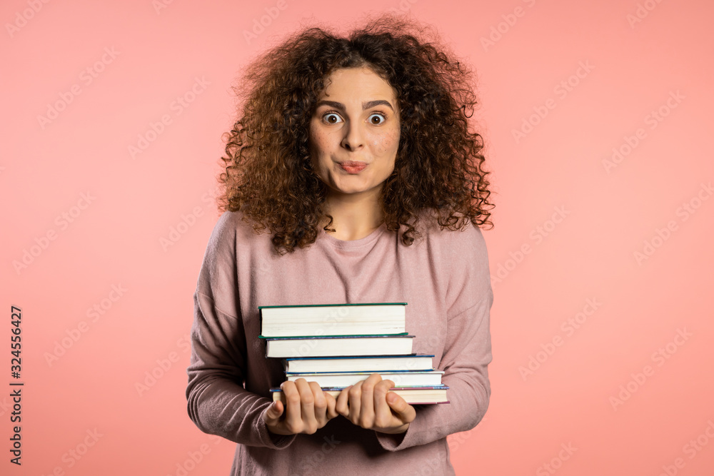 European student on pink background in studio holds stack of university books from library. Woman is happy to graduate. European student on pink background in studio holds stack of university books from library. Woman is happy to graduate.