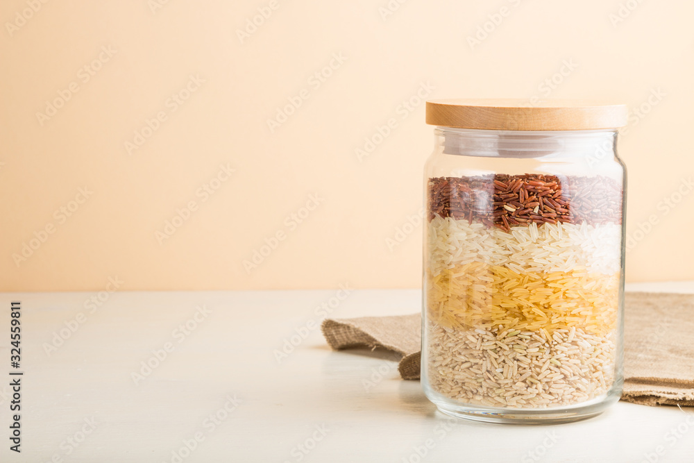 glass jar with different kinds of rice poured in layers on white and orange background. side view, copy space. glass jar with different kinds of rice poured in layers on white and orange background. side view, copy space.
