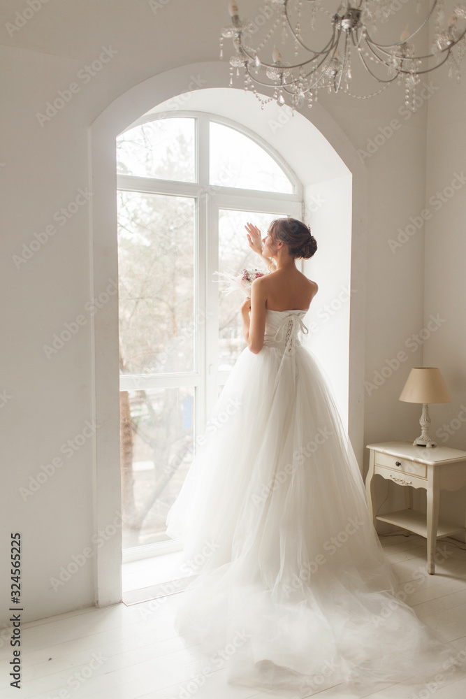 Fototapeta premium Young bride in a beautiful dress holding a bouquet of flowers posing near window in bright white studio. Wedding concept.