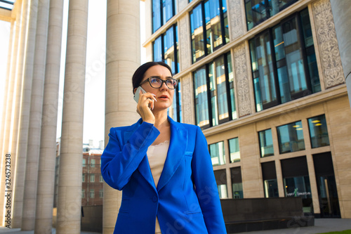 Serious woman in glasses and official wear successful government worker talking via mobile phone while standing outdoors near enterprise during work break. Female lawyer having cellphone conversation