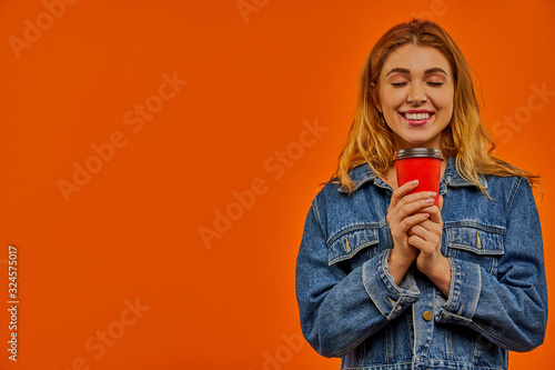 A woman with wavy fair hair in a denim jacket smiles with closed eyes and holds a red cardboard cup of coffee in her hands.