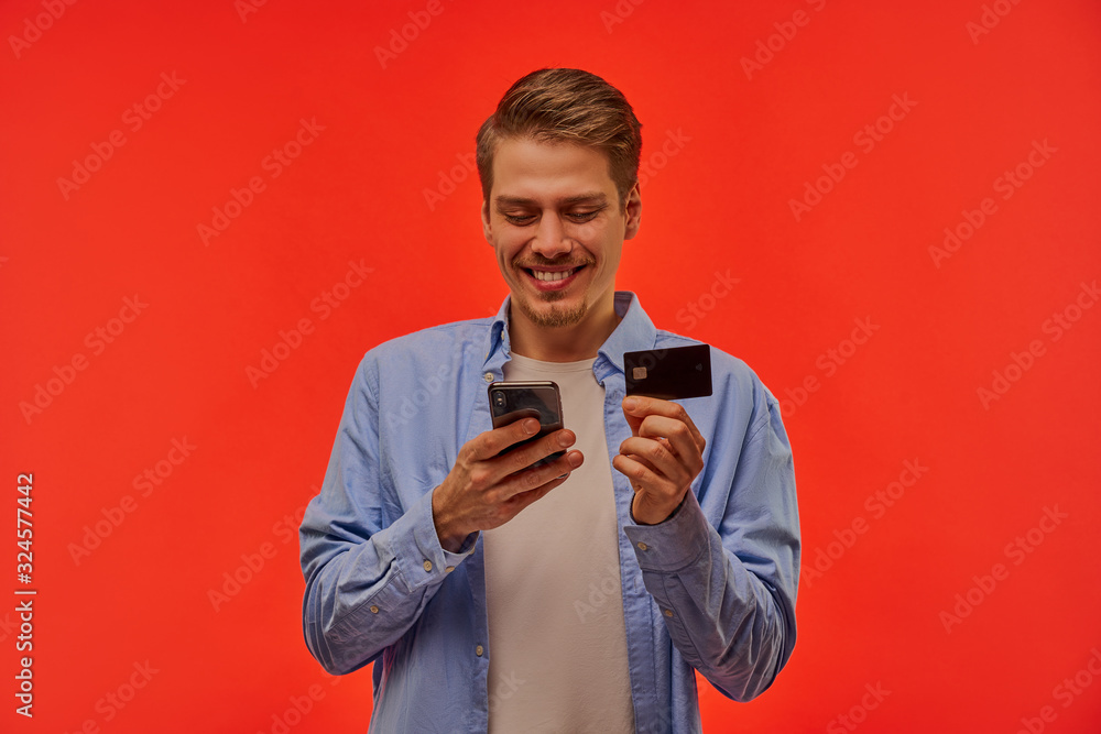 Happy man with a beard in a blue shirt looks into phone and shows a black credit card. Happy man with a beard in a blue shirt looks into phone and shows a black credit card.