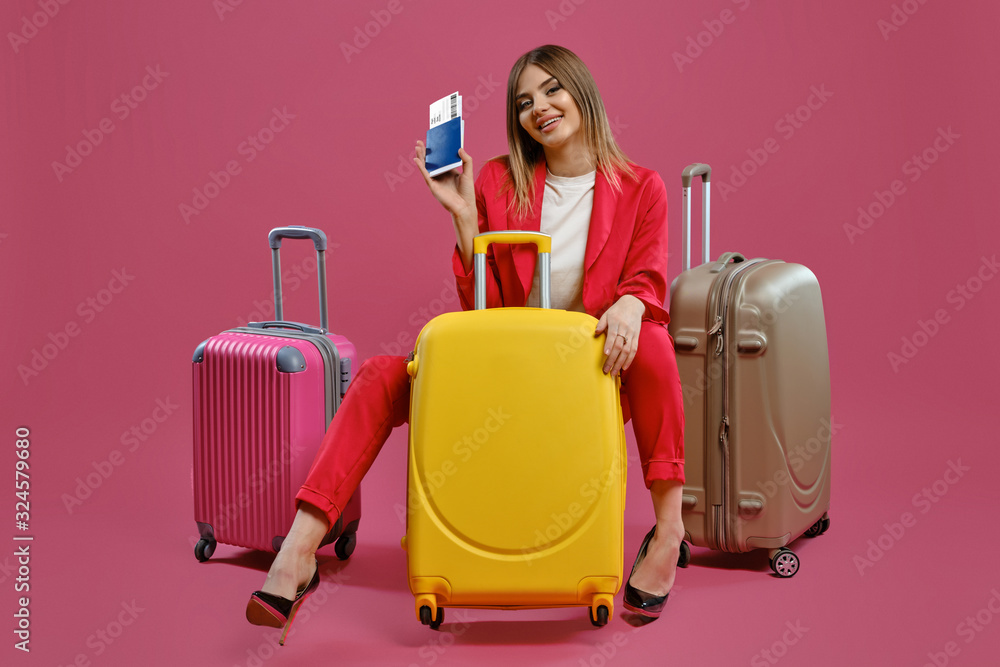 Blonde model in red pantsuit, white blouse, black heels. She smiling, sitting among colorful suitcases, holding passport and ticket, pink background Blonde model in red pantsuit, white blouse, black heels. She smiling, sitting among colorful suitcases, holding passport and ticket, pink background