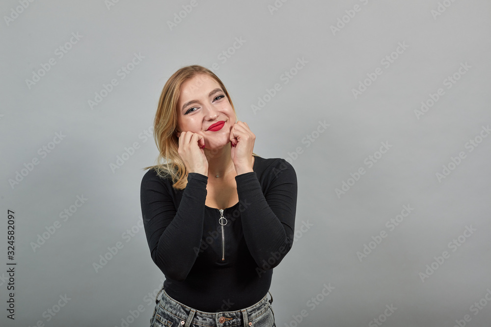 Young blonde girl in black jacket on grey background happy woman touches her face with hands Young blonde girl in black jacket on grey background happy woman touches her face with hands