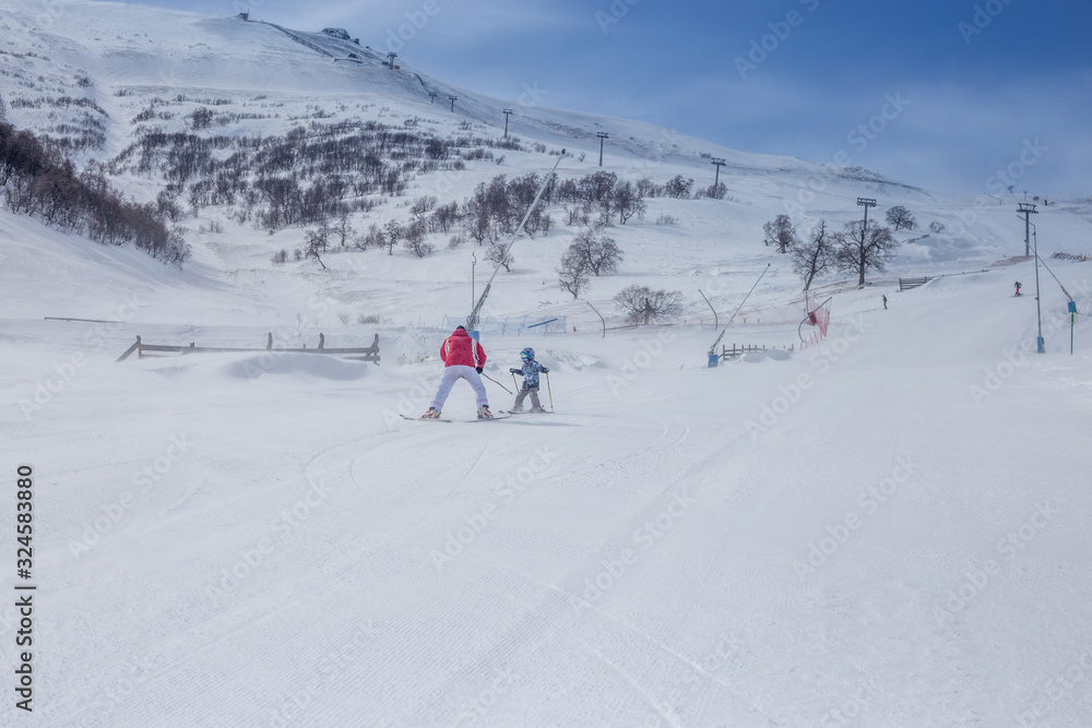 A small child learns to ski. A girl in a blue jacket and helmet rides down the slope to the instructor in a red jacket. Georgia, Bakuriani, Caucasus.