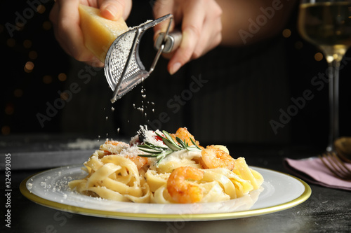Fotografie Woman grating cheese onto pasta with shrimps on black table, closeup