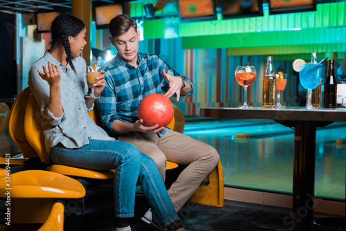 young man showing bowling ball to smiling african american girl holding cocktail