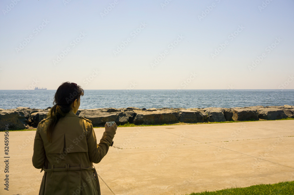 Obraz premium Young sad woman standing alone near at seaside outdoor, drinking tea / coffee, looking at distant seascape horizon. Copy space for text.