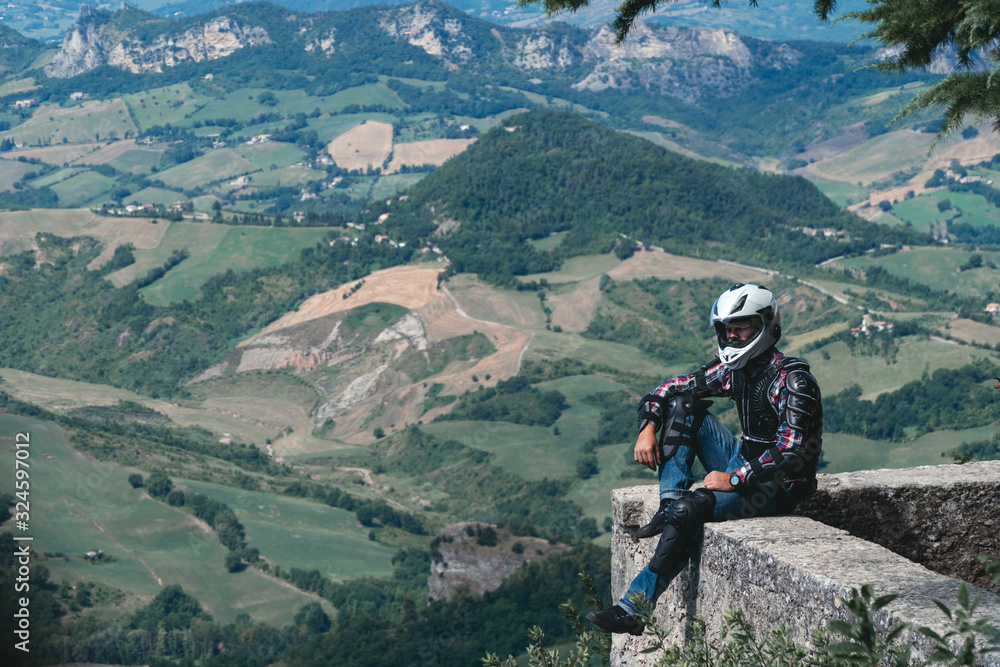 Man dressed in a motorcycle outfit. body protection turtle and knee pads. Biker is sitting on the edge of the top of the mountain. incredible view. Motorcycle travel concept Copy space. San Marino