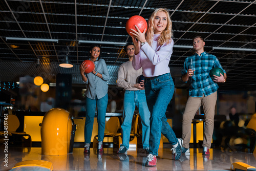 Billede på lærred smiling blonde girl throwing bowling ball near multicultural friends