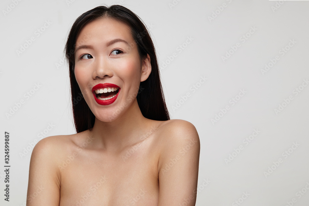 Joyful young lovely dark haired lady with festive makeup smiling happily while looking excitedly aside, keeping hands down while standing over white background Joyful young lovely dark haired lady with festive makeup smiling happily while looking excitedly aside, keeping hands down while standing over white background