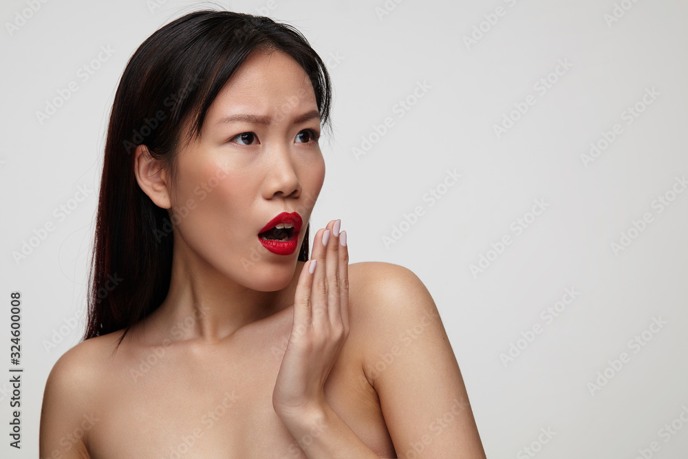 Amazed young attractive dark haired lady with festive makeup raising hand to her mouth while looking surprisedly aside, standing against white background Amazed young attractive dark haired lady with festive makeup raising hand to her mouth while looking surprisedly aside, standing against white background