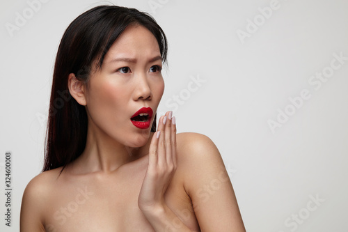 Amazed young attractive dark haired lady with festive makeup raising hand to her mouth while looking surprisedly aside, standing against white background