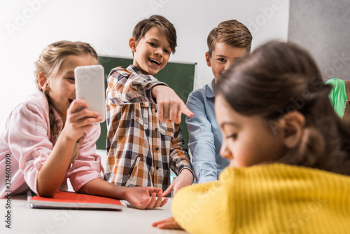 selective focus of schoolkid with smartphone and cruel classmates laughing near bullied kid, cyberbullying concept photo