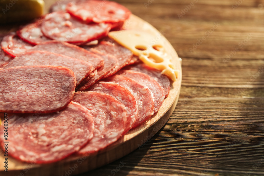 Thin slices of sausage, cheese and pickled cucumbers on a round cutting board on a wooden table. Russian appetizer. Thin slices of sausage, cheese and pickled cucumbers on a round cutting board on a wooden table. Russian appetizer.
