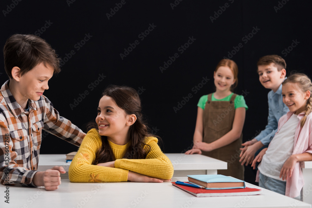 selective focus of schoolboy smiling while looking at schoolgirl near classmates isolated on black selective focus of schoolboy smiling while looking at schoolgirl near classmates isolated on black