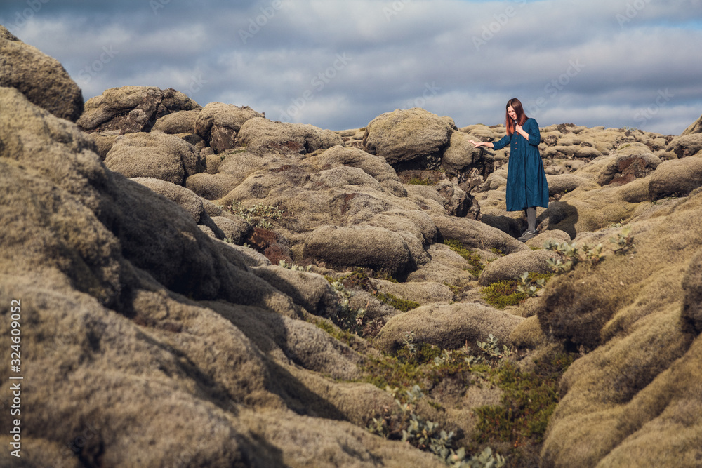 Young beautiful woman in blue dress stands on the volcanic formations covered by green moss on a background sky. Place for text or advertising