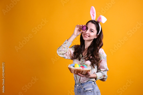 Studio portrait of young beautiful woman wearing traditional bunny ears headband for easter and smiling. Brunette female with wavy hair over yellow background. Close up, copy space.