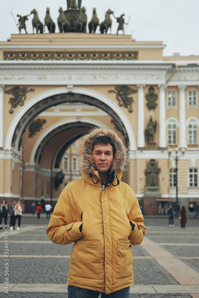 Young stylish man tourist walking near General Staff Building arch in the palace square in St. Petersburg. He wearing in yellow winter jacket