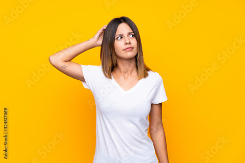 Young woman over isolated yellow background having doubts while scratching head