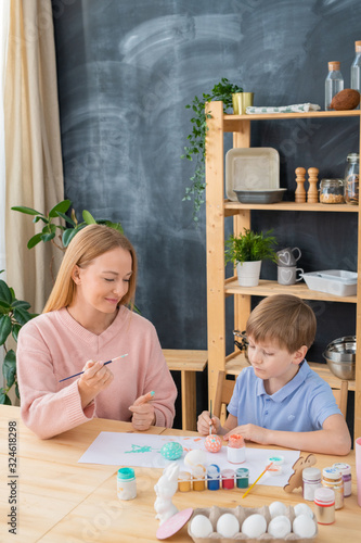 Serious boy sitting at table with papers and painting eggs while creating Easter design with mother