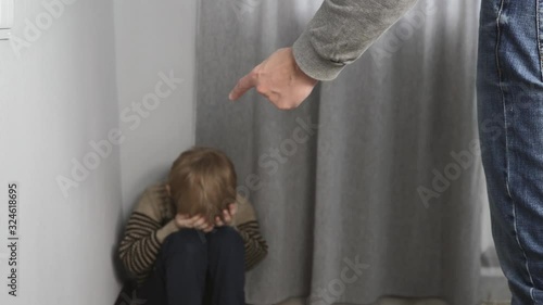 A frightened, crying child is sitting in the corner of the room. Violence against children in the family. The male father holds the belt in his hand.