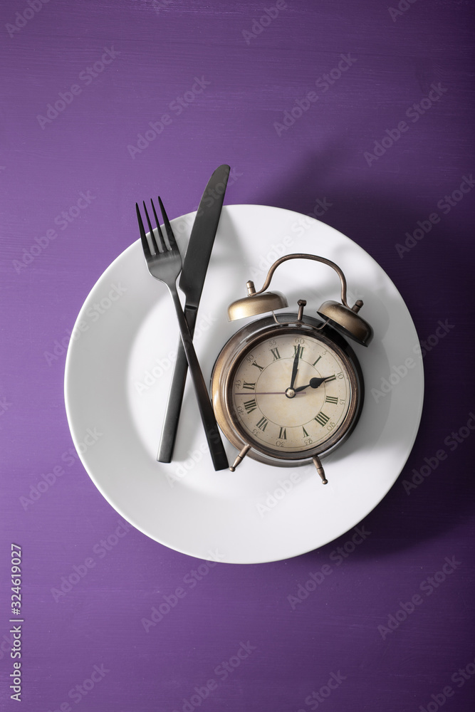 concept of intermittent fasting, ketogenic diet, weight loss. fork and knife crossed on a plate and alarmclock concept of intermittent fasting, ketogenic diet, weight loss. fork and knife crossed on a plate and alarmclock