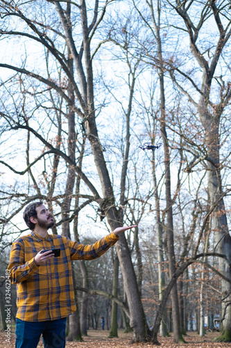 Young man flying a drone with a controller in a park with some trees and sky in background.