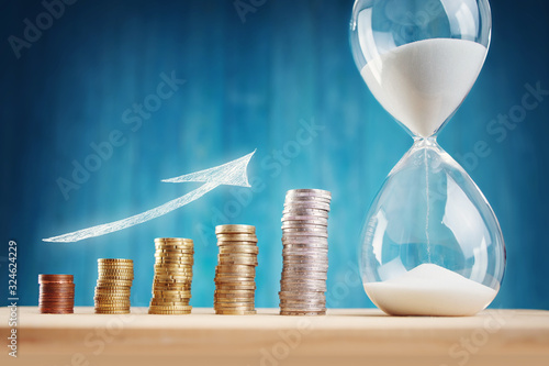 clock with stacked coins and hourglass on blue background photo