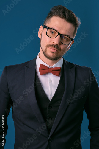 Young business man in classic black suit, white shirt, red bow-tie and glasses. Posing against blue studio background. Mock up, copy space. Close-up. © nazarovsergey