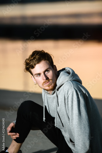 Young redhead man posing at sunset next to a pier dressed casual wearing gray sweatshirt.