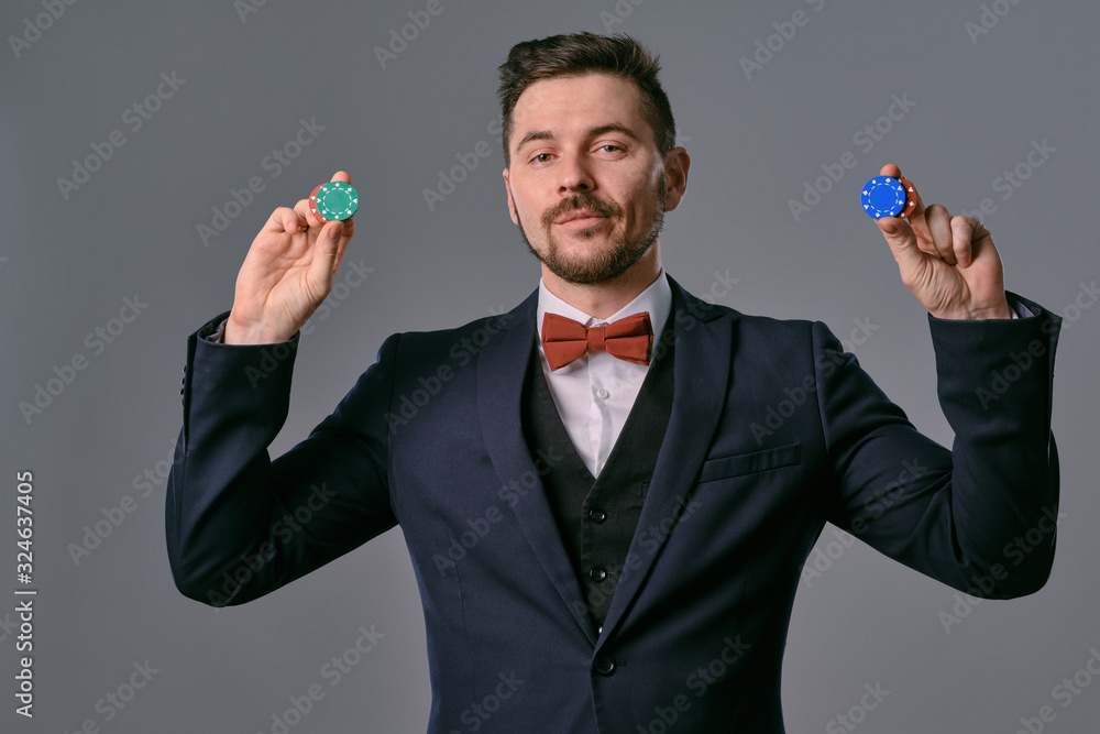 Fototapeta premium Man in black classic suit and red bow-tie showing some colored chips, posing against gray studio background. Gambling, poker, casino. Close-up.