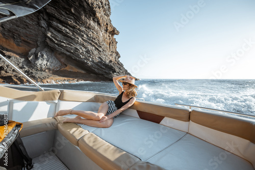 Relaxed attractive woman in swimsuit and sun hat enjoying ocean voyage, sailing on the luxury yacht near the beautiful rocky coast on a sunset. Concept of a summer luxury rest on the sea