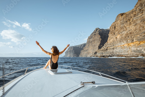 Woman enjoying ocean voyage sitting back on the yacht nose while sailing near the breathtaking rocky coast on a sunset