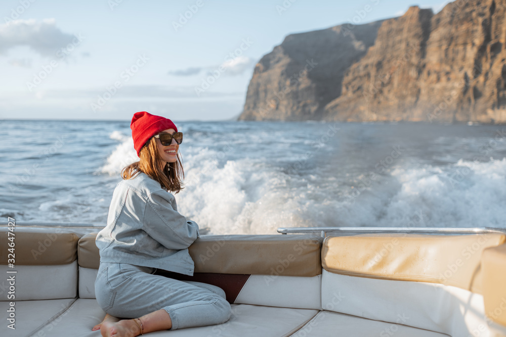 Portrait of a young woman traveler dressed casually in red hat and jeans sailing on a yacht near the rocky coast. Concept of a carefree lifestyle and travel
