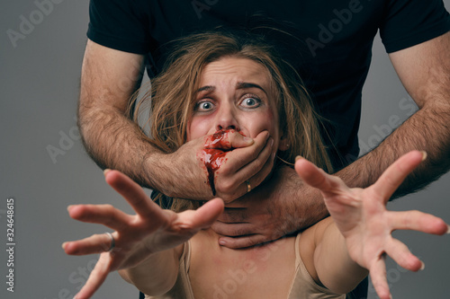 Strong man strangling scared female with bruises on her face. Blood from her mouth flows down his arm. Gray background. Domestic violence. Close-up. photo