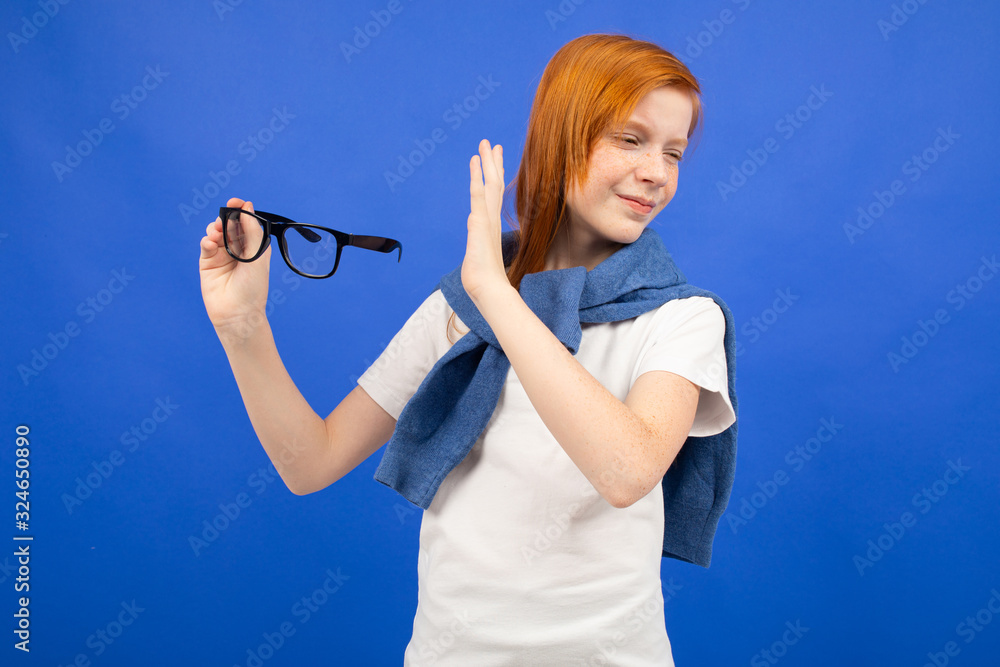 red-haired teenage girl in a white T-shirt throws out glasses on a blue studio background. new vision red-haired teenage girl in a white T-shirt throws out glasses on a blue studio background. new vision