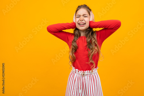 cute teenager girl with curly hair in a red blouse and striped trousers squints and closed her ears on a yellow background with copy space