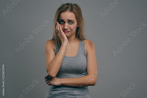 Blonde girl with bruises is touching her face her face, gray studio background. Domestic violence, abuse. Depression, despair. Close-up, copy space.