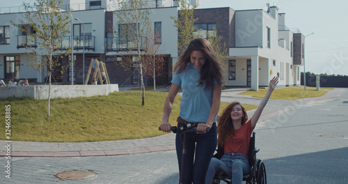 Appealing girl riding electric powered scooter pulling along a ginger curly girl in wheelchair spending fun spare time together in modern city on sunny day. photo