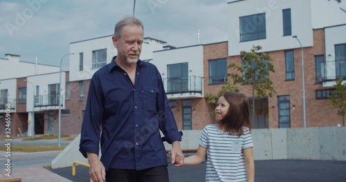 Cheerful grandfather walking with beautiful cute lgirl kid in the street talking together spending spare time outdoors in the neighbourhood. photo