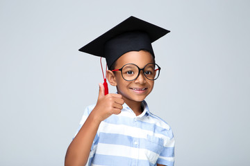 Young African American boy in graduation cap showing thumb up on grey background