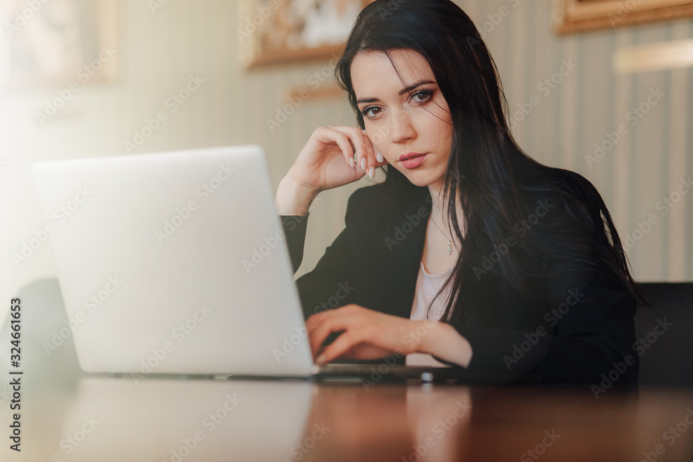 Young attractive emotional girl in business-style clothes sitting at a desk on a laptop and phone in the office or auditorium Young attractive emotional girl in business-style clothes sitting at a desk on a laptop and phone in the office or auditorium