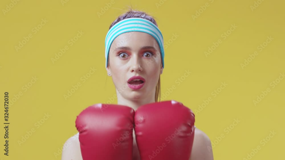 Beautiful fitness woman kickboxer appearing behind red boxing gloves gives a strong feminine look hitting camera posing on yellow background. Strong woman concept. Beautiful fitness woman kickboxer appearing behind red boxing gloves gives a strong feminine look hitting camera posing on yellow background. Strong woman concept.