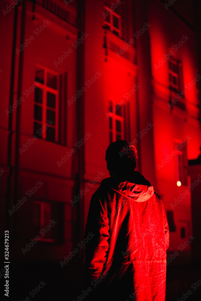 Back view silhouette of man standing by building illuminated in red looking in windows at night
