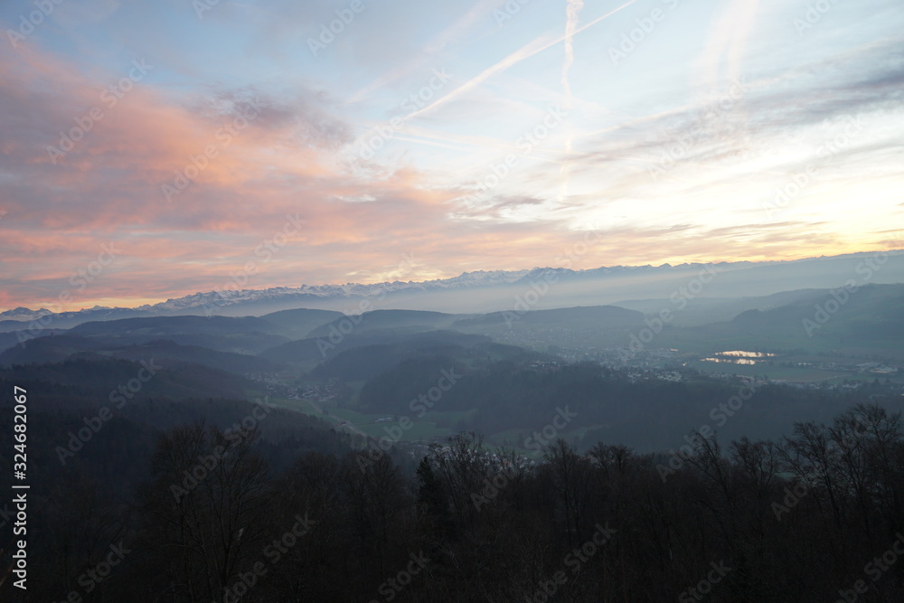 Sunset panorama of Alps over canton Zurich, Switzerland. The whole landscape is veiled in fog. The sky is colorful at sunset. There is a valley among moderate hills and two small lakes.