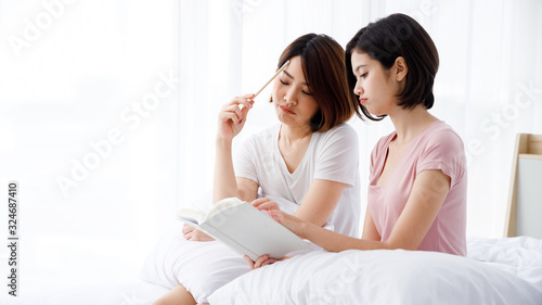 Two Asian girl sit on bed and study or do homework together in the morning. Pink shirt woman is point at notebook and the other one is thinking.