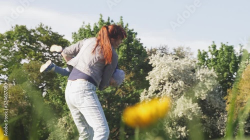 Pretty woman is circling baby boy dressed in blue jacket on her hands and having fun with kid in spring sunny city park with blooming trees on background. 4K slow motion video.