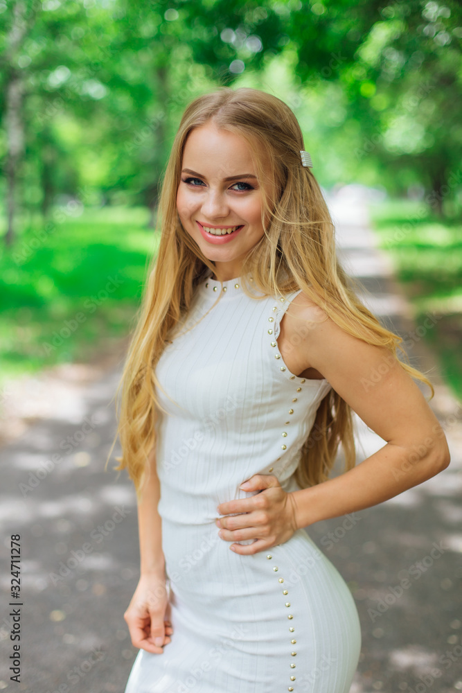 Portrait of a charming blond woman wearing beautiful white dress standing on the road under trees.