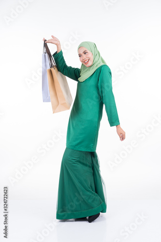 A beautiful Asian female model in a modern kurung carrying shopping bags isolated on white background. Eidul fitri festive preparation shopping concept. Full length portrait. photo